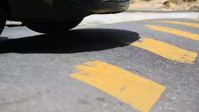 A vehicle drives over a yellow-painted speed bump on a paved road.